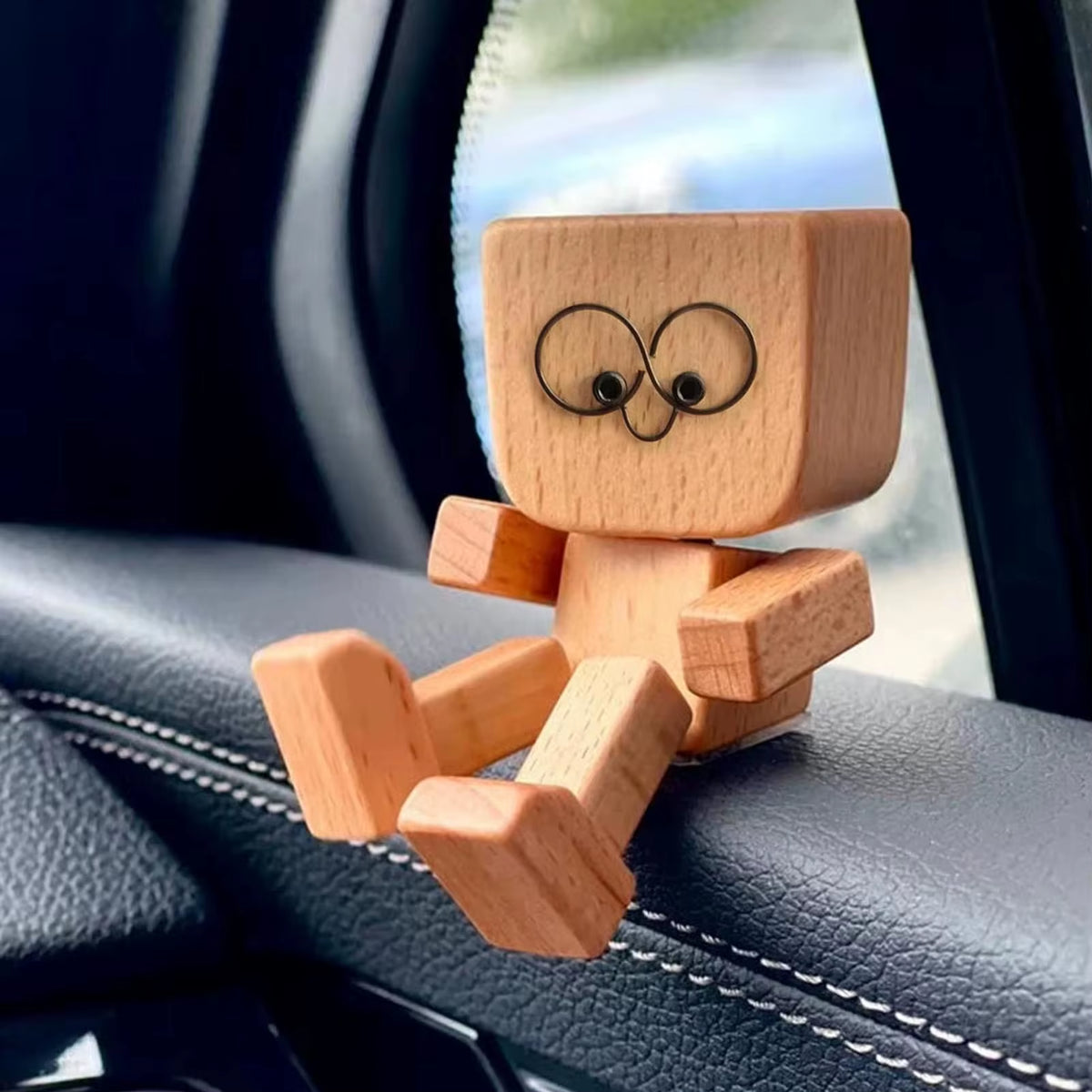 Wooden figurine seated on a car dashboard near a seatbelt, with wire-loop eyes and a small screw nose
