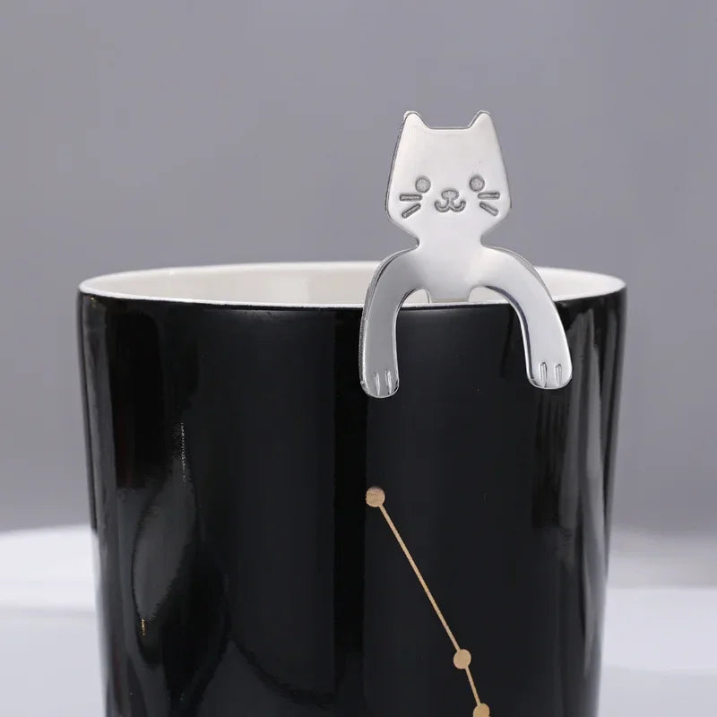 Close-up of a silver cat spoon hooked onto the rim of a black mug with a gold constellation design