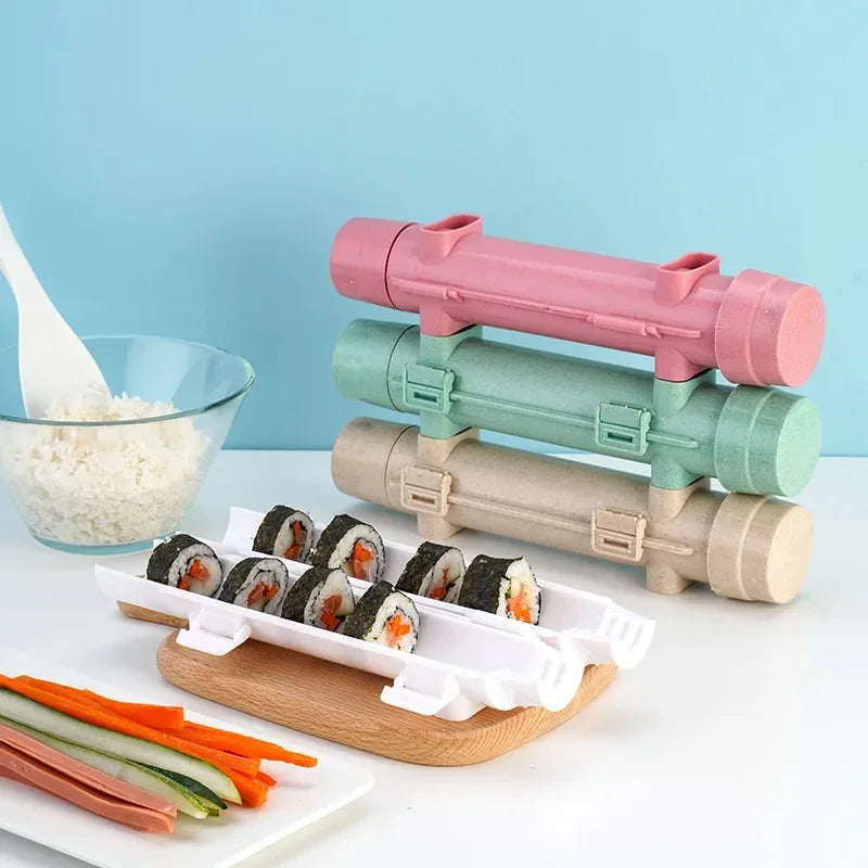 Pink, green, and beige sushi tubes stacked pyramid-style beside a nori tray, rice bowl, and veggie strips on blue