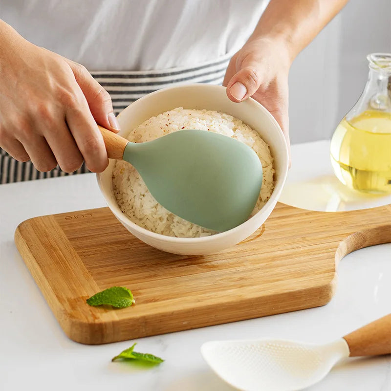 Hands using the sage green paddle to scoop rice into a bowl on a wood cutting board with mint leaves