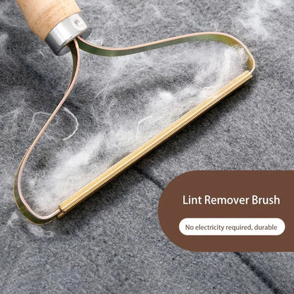 Angled shot of the comb with a thick clump of white lint caught in the gold teeth against grey fabric, labeled Lint Remover Brush