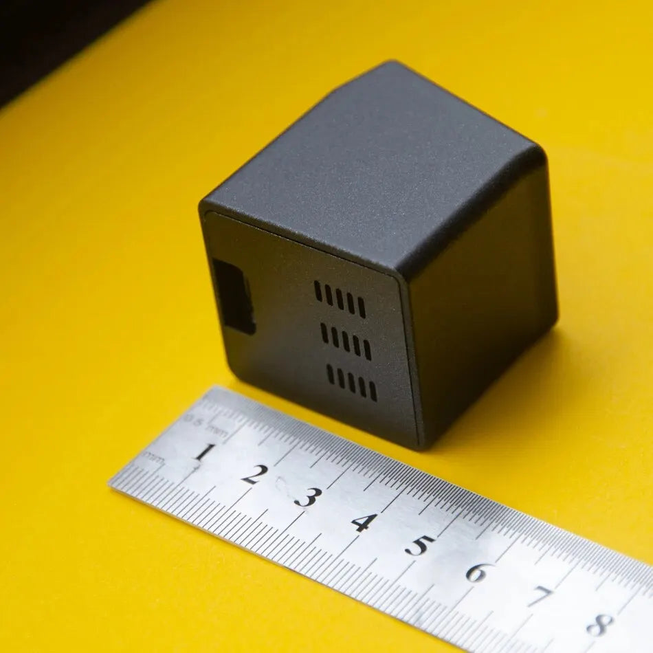Rear view of black weather clock beside a metal ruler on yellow background showing its compact size