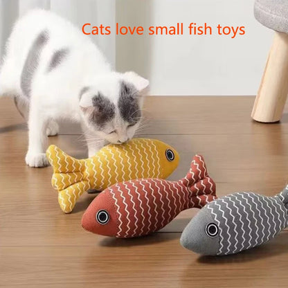 White and grey kitten sniffing yellow, red, and grey striped fabric fish toys on a wood floor, text reads cats love small fish toys