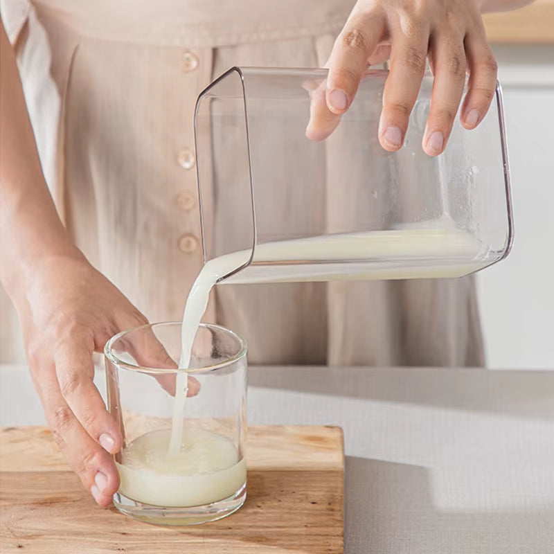 Hands pouring milk from a clear pitcher into a glass on a wood board