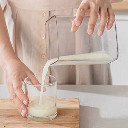Hands pouring milk from a clear pitcher into a glass on a wood board