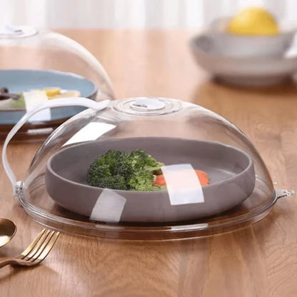 Two splash guard domes on a wood table, one over a gray bowl of broccoli and carrot, another over a blue plate, lemon nearby