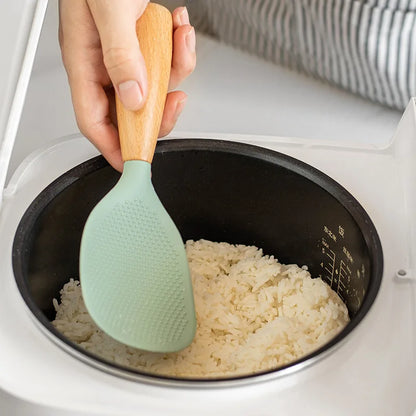 Hand using the sage green silicone paddle to scoop rice from a black electric rice cooker