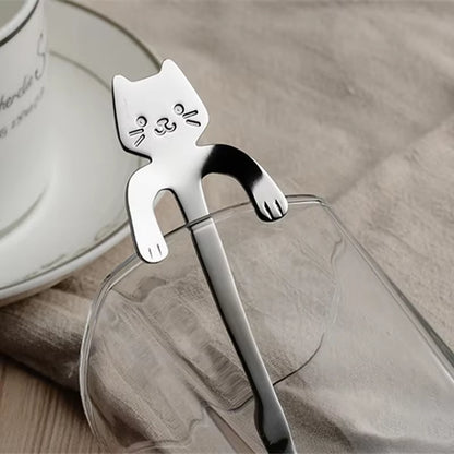 Close-up of a silver cat spoon hooked onto the edge of a glass, teacup blurred in the background