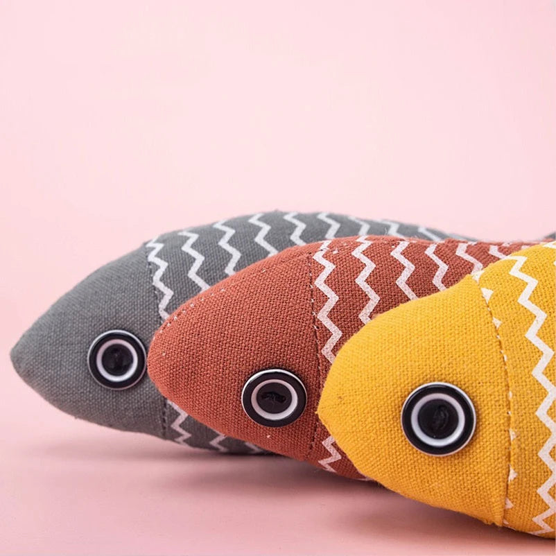 Close-up of three striped fabric fish toys in grey, rust, and yellow with button eyes, on pink background