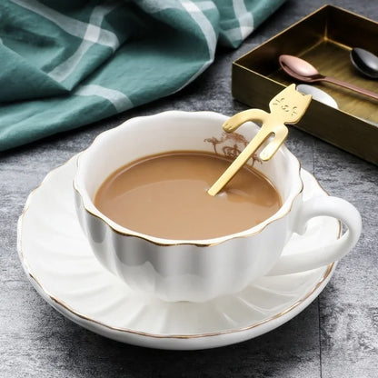 Gold cat spoon hooked on a teacup of coffee, with green cloth and a tray of rose gold and black spoons